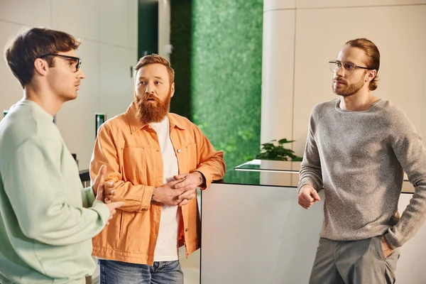 Thoughtful bearded man in stylish casual clothes looking at serious business partners discussing business project in hall of modern office space, brainstorming, startup planning, problem-solving — Stock Photo