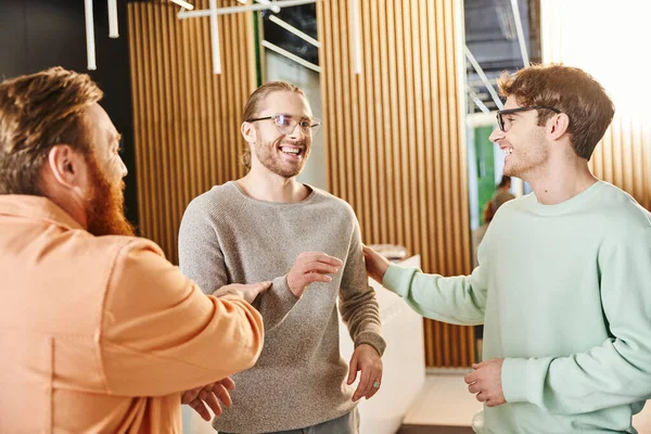 Empreendedores alegres em óculos e roupas casuais elegantes discutindo projeto de startup perto parceiro de negócios barbudo no salão do espaço de co-trabalho contemporâneo, brainstorming e colaboração — Fotografia de Stock