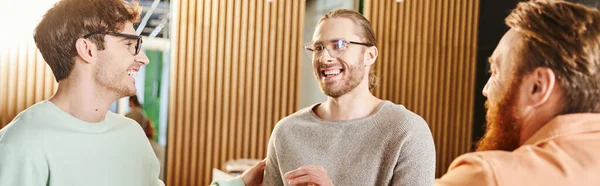 Stylish and cheerful businessmen in eyeglasses smiling during discussion of startup project near bearded colleague in modern coworking space, partnership of successful entrepreneurs, banner — Stock Photo