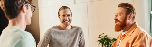 Homem de negócios barbudo alegre que sorri perto de colegas inspirados em óculos que falam durante o encontro no corredor do espaço de coworking contemporâneo, parceria e colaboração, bandeira — Fotografia de Stock