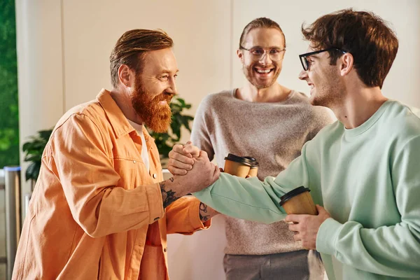 Empresarios llenos de alegría con bebidas para llevar estrechando las manos cerca de colega sonriente en gafas, socios de negocios exitosos cerrando trato durante el descanso de café en la oficina contemporánea - foto de stock