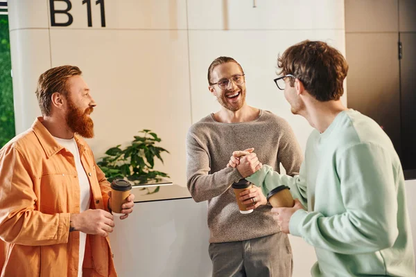 Excited business partners in eyeglasses shaking hands as gesture of agreement near bearded colleague, entrepreneurs with takeaway coffee in paper cups meeting in lobby of modern coworking space — Stock Photo