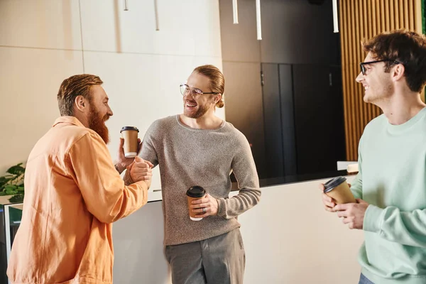 Cheerful businessmen with takeaway drinks in paper cups shaking hands near happy colleague, successful entrepreneurs closing deal during coffee break in hall of modern coworking office — Stock Photo