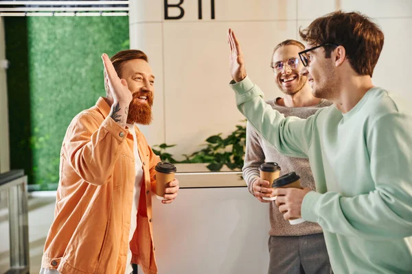 Cheerful business partners with paper cups confirming agreement and giving high five near pleased colleague in eyeglasses during coffee break in hall of modern coworking space — Stock Photo