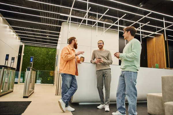 Бородатый мужчина держит бумажный стаканчик и разговаривает с улыбающимися коллегами в вестибюле современного коворкинга, встречается с успешными бизнесменами, обсуждающими стартап-проект во время кофе-брейка — стоковое фото