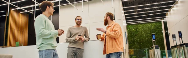 Оптимістичний бородатий бізнесмен жестикулює і розмовляє зі усміхненими колегами в сучасному офісному просторі, спілкування стильних підприємців під час зустрічі на кавовій перерві, банер — стокове фото