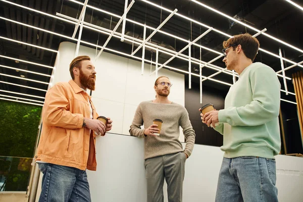 Low angle view of businessmen in stylish casual clothes holding takeaway drinks and discussing startup project during meeting at coffee break in modern coworking office with high tech interior — Stock Photo