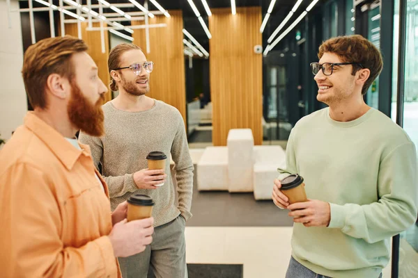 Bearded man with stylish and cheerful colleagues in eyeglasses holding takeaway coffee during conversation in lobby of modern coworking space, ambitious businessmen discussing startup project — Stock Photo