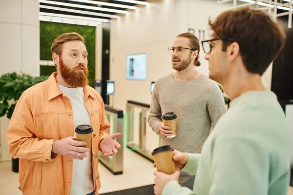 Серьезный бородатый мужчина разговаривает с коллегами в очках держа бумажные стаканчики с напитками на вынос во время кофе-брейка в современном коворкинг-офисе, партнерства и сотрудничества в современном бизнесе — стоковое фото