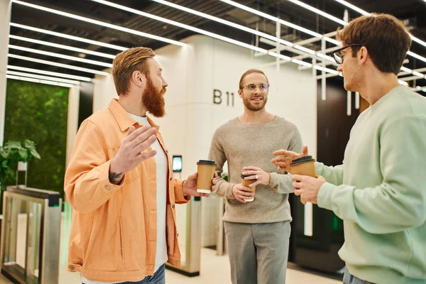 Positive entrepreneurs holding paper cups with coffee to go during conversation in lobby of contemporary office, businessmen discussing startup project, collaboration and productivity — Stock Photo
