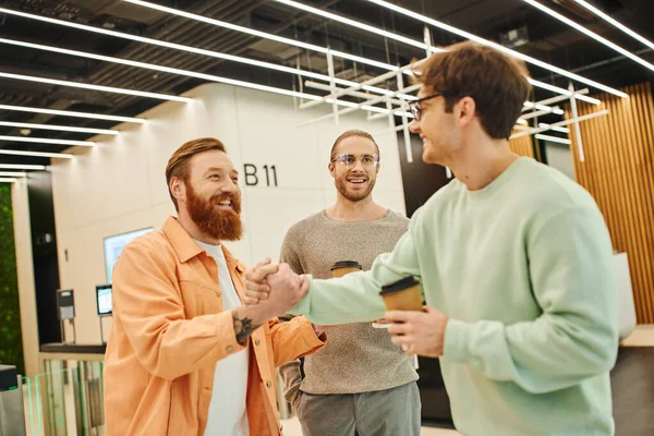 Щасливий бородатий чоловік тремтить руками з бізнесменом в окулярах біля усміхненого колеги, бізнес-партнери з кавою, щоб піти на угоду в лобі сучасного коворкінгу — стокове фото