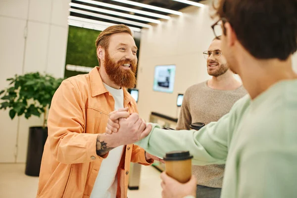 Happy bearded entrepreneur shaking hands with business partner near smiling colleague while holding paper cups during coffee break in lobby of modern office space, success and cooperation concept — Stock Photo
