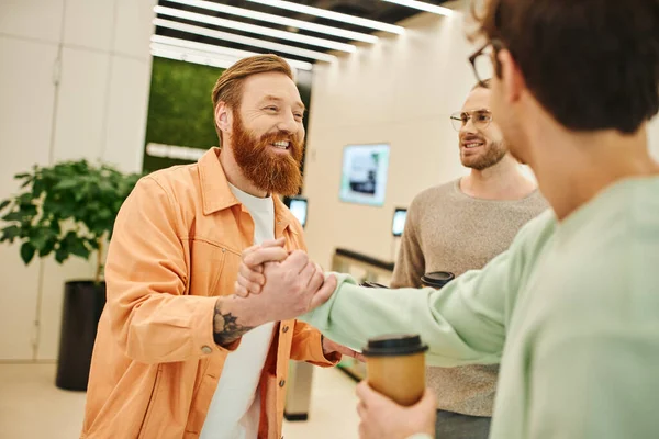 Empreendedor barbudo feliz apertando as mãos com parceiro de negócios perto de colega sorridente enquanto segurando copos de papel durante pausa para café no lobby do moderno espaço de escritório, sucesso e conceito de cooperação — Fotografia de Stock