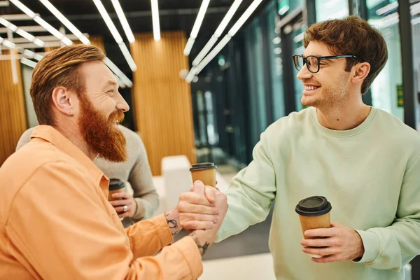 Happy bearded man shaking hands with business partner in eyeglasses near colleague on blurred background, pleased entrepreneurs with paper cups confirming agreement during coffee break — Stock Photo