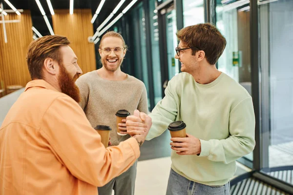 Overjoyed businessmen holding paper cups with takeaway drink and shaking hands near colleague in contemporary coworking environment, successful entrepreneurs closing deal during coffee break — Stock Photo
