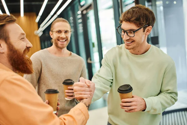 Pleased bearded man and his partner in eyeglasses holding takeaway drinks and shaking hands near colleague in hall of modern office, successful entrepreneurs closing deal during coffee break — Stock Photo