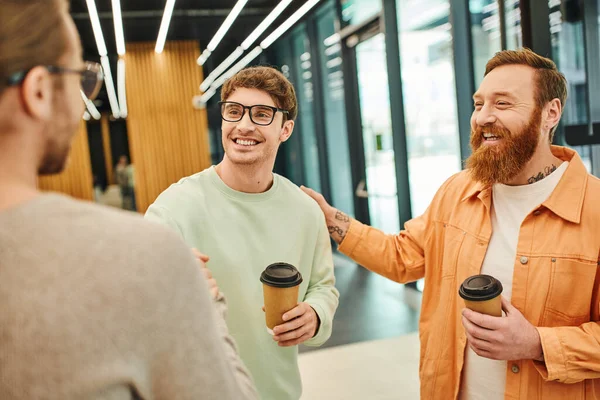 Позитивный бородатый мужчина трогает плечо счастливого коллеги пожимающего руку бизнесмену на размытом переднем плане, успешные предприниматели с кофе идут на подтверждение соглашения в современном офисе — стоковое фото