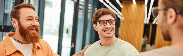 Joyous business partners talking and looking at each other in modern coworking office, tattooed bearded man with colleagues in eyeglasses, successful collaboration, blurred foreground, banner — Stock Photo
