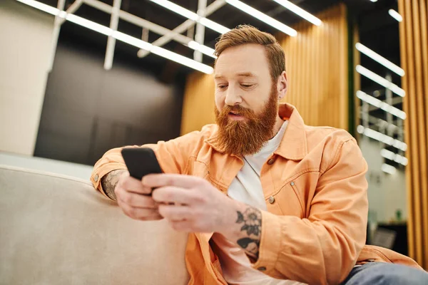 Татуйований, бородатий і стильний підприємець в повсякденному одязі, сидячи на зручному дивані в залі сучасного коворкінгу і переглядаючи інтернет на мобільному телефоні — стокове фото