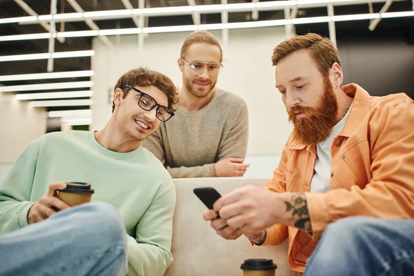 Homens de negócios sorridentes em óculos olhando para redes de colegas barbudos e tatuados, usando telefone celular enquanto sentados no sofá no salão do espaço contemporâneo de co-trabalho durante o coffee break — Fotografia de Stock