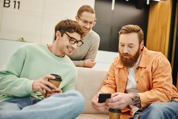 Happy businessmen in eyeglasses looking at bearded colleague browsing internet on mobile phone on comfortable couch, coffee break in lounge of modern coworking space — Stock Photo