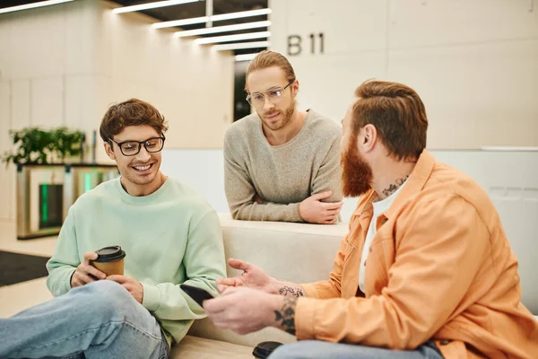 Бородатий чоловік з мобільним телефоном розмовляє зі усміхненими колегами в окулярах, сидячи на зручному дивані під час кавової перерви в залі сучасного офісу, позитивні підприємці обговорюють стартап — стокове фото