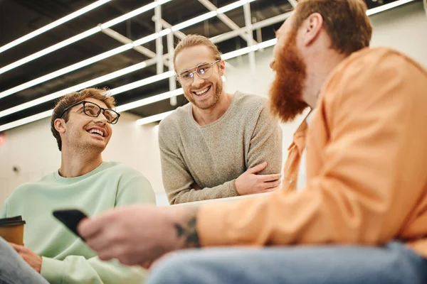 Low angle view of bearded entrepreneur with mobile phone discussing possibility of business startup with cheerful colleagues in eyeglasses during coffee break in modern office on blurred foreground — Stock Photo
