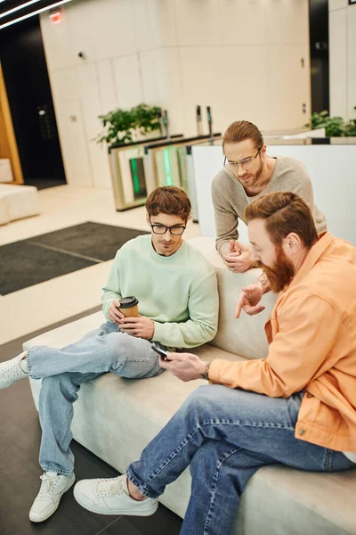 Bearded businessmen pointing with finger and showing smartphone to colleagues in eyeglasses while sitting on couch in lounge of modern coworking office during coffee break — Stock Photo