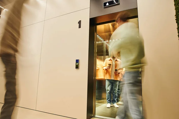 Desenfoque movimiento de los hombres de negocios cerca de colega sosteniendo tazas de papel con café para llevar mientras está de pie en el ascensor, pausa para el café en la oficina contemporánea de coworking, interior de alta tecnología, lugar de trabajo moderno - foto de stock