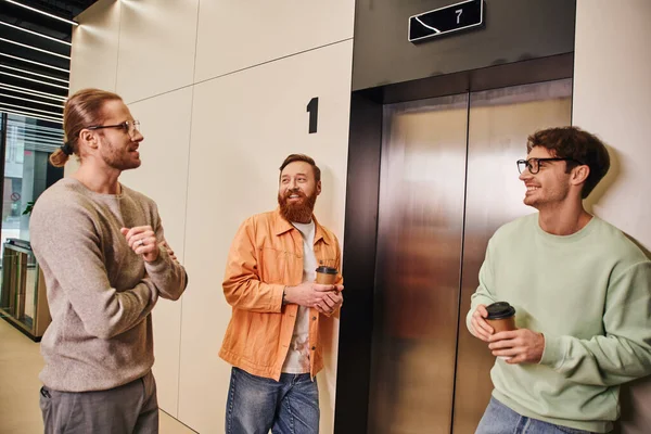 Zufriedene Geschäftskollegen in stylischer Freizeitkleidung mit Imbissgetränken in Pappbechern beim Warten auf den Fahrstuhl, erfolgreiche Unternehmer, die sich in der Kaffeepause im modernen Büro unterhalten — Stockfoto