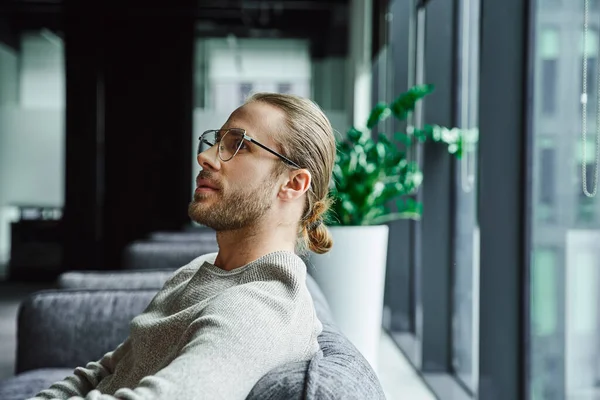 Seitenansicht eines nachdenklichen Geschäftsmannes in stylischer Brille und lässiger Kleidung, der an ein Startup-Projekt denkt, während er in einer Bürolounge in zeitgenössischem Coworking Space sitzt, Ehrgeiz und kreatives Denken — Stockfoto