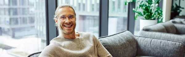 Professional headshot of smiling and successful businessman in casual clothes and stylish eyeglasses sitting on comfortable couch in lounge of modern coworking office and looking at camera, banner — Stock Photo