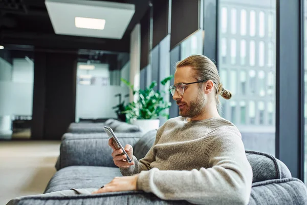 Вид сбоку серьезного и стильного бизнесмена в повседневной одежде и очках, смотрящего в смартфон, сидя на удобном диване в окружении современной офисной среды — стоковое фото
