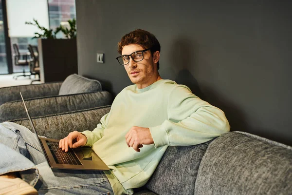 Entrepreneur ambitieux dans des lunettes élégantes et des vêtements décontractés assis sur un canapé confortable avec ordinateur portable et regardant la caméra dans l'environnement de bureau contemporain, la confiance et le concept de succès — Photo de stock
