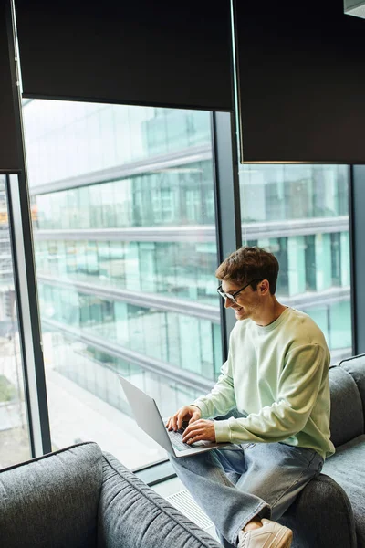 Joyful businessman in casual clothes and eyeglasses typing on laptop while sitting on comfortable couch near large windows in lounge of coworking office, modern workplace, business productivity — Stock Photo