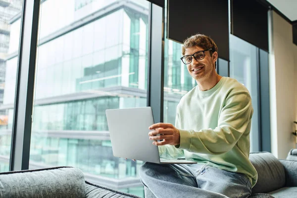 Energetic and overjoyed businessman in stylish eyeglasses sitting near large windows in lounge of modern coworking space and smiling at camera, business lifestyle, ambition and success — Stock Photo