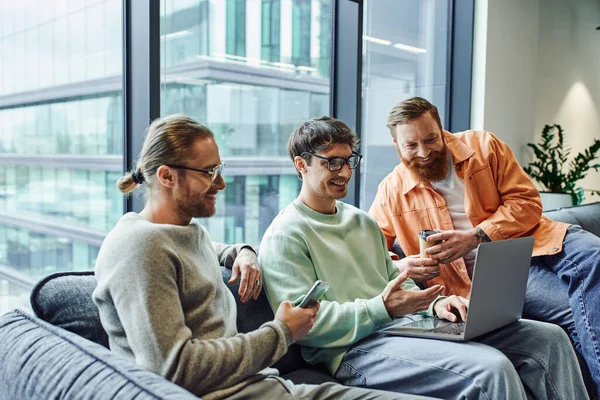 Happy business colleagues with mobile phone, laptop and coffee to go sitting on comfortable sofa, successful entrepreneurs networking in lounge of contemporary coworking environment — Stock Photo