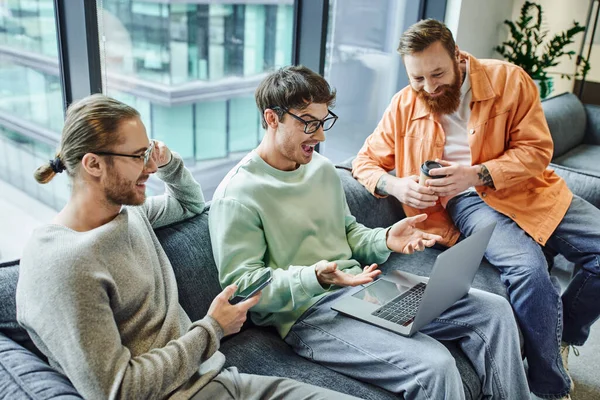 Excited entrepreneur with open mouth pointing at laptop near happy business partners with paper cup and mobile phone on couch in lounge of contemporary coworking office, partnership and success — Stock Photo