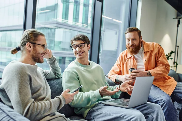 Happy businessman in eyeglasses pointing at laptop near pleased colleagues with smartphone and coffee to go, office lounge in modern coworking environment, collaboration, successful startup project — Stock Photo