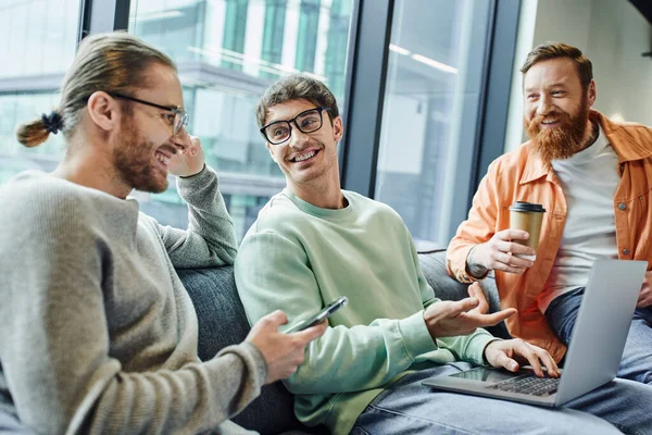 Satisfied entrepreneur in eyeglasses pointing at laptop while working on business planning near colleagues with smartphone and coffee to go on couch in lounge of modern office, coworking and success — Stock Photo