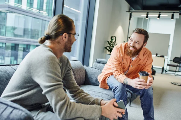 Cheerful, bearded and tattooed businessman with coffee to go discussing startup project with colleague sitting with mobile phone on couch in office lounge of modern coworking environment — Stock Photo