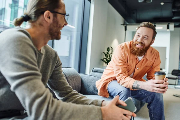 Blurred businessman holding smartphone while talking to happy and bearded colleague sitting with takeaway drink, discussing and planning startup on sofa in contemporary coworking office — Stock Photo