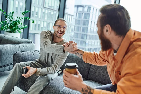 Joyful entrepreneur in eyeglasses sitting on couch with smartphone and shaking hands with bearded businessman with paper cup on blurred foreground, confirming agreement in modern office — Stock Photo