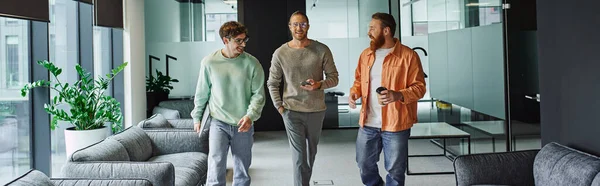 Bearded entrepreneur with coffee to go talking to joyful and stylish business partners walking with smartphone and laptop in contemporary coworking environment and planning startup, banner — Stock Photo
