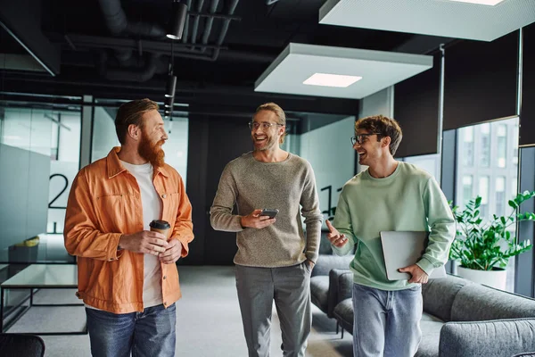 Excited businessmen in eyeglasses walking with laptop and mobile phone and talking to bearded colleague in contemporary coworking space, partnership and teamwork in startup project — Stock Photo