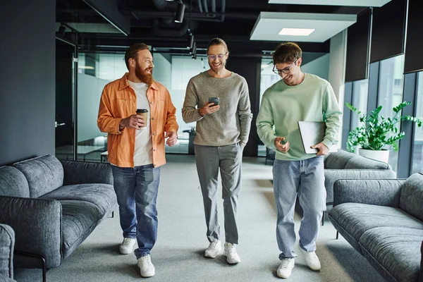 Cheerful entrepreneurs in stylish casual clothes walking with mobile phone, laptop and takeaway drink in comfortable lounge of modern office and discussing collaboration in business project — Stock Photo
