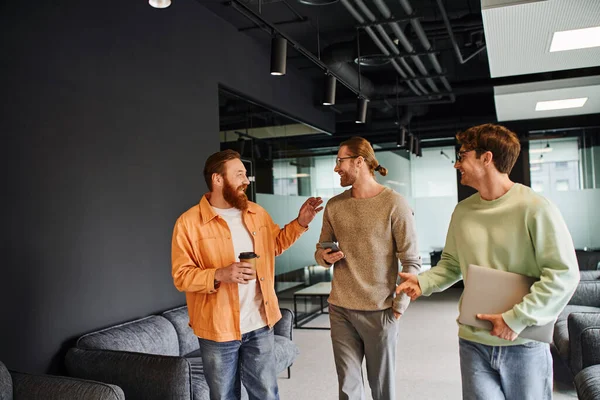 Joyous bearded entrepreneur holding coffee to go and talking to business partners with smartphone and laptop walking in contemporary lounge of coworking space, successful collaboration — Stock Photo