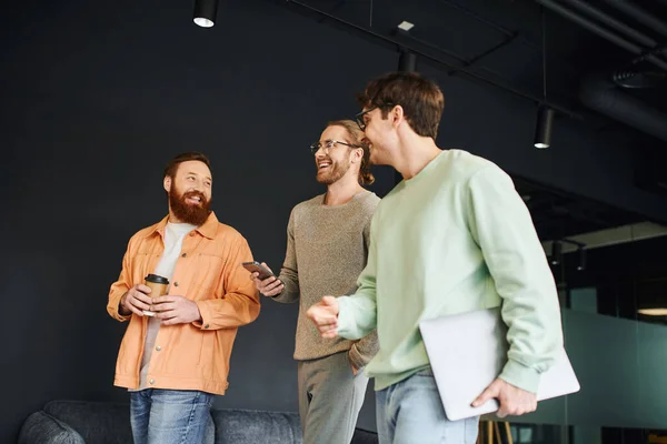 Overjoyed bearded man with takeaway drink in paper cup looking at colleagues with smartphone and laptop walking in modern coworking office and discussing business project — Stock Photo