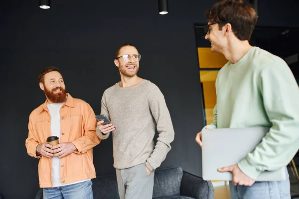 Cheerful entrepreneurs in stylish casual clothes, with mobile phone and takeaway drink, looking at colleague with laptop on blurred foreground in modern coworking space, successful partnership — Stock Photo