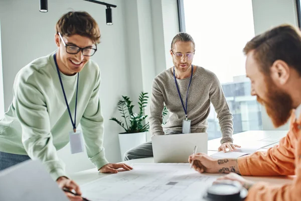 Smiling architect pointing at blueprint near colleagues and laptop while working in coworking space of architectural studio, business collaboration and implementation of innovative ideas — Stock Photo